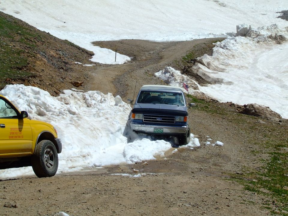 1987 Ford Bronco driving through snow on a dirt road, yellow truck to the left