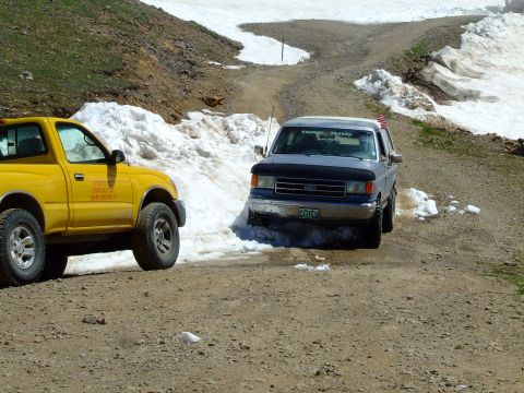1987 Ford Bronco - A blue Ford Bronco navigates a snowy mountain road, accompanied by a yellow pickup truck.