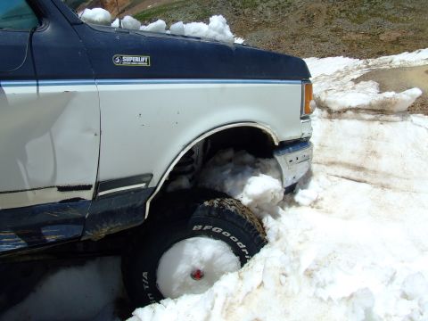 1987 Ford Bronco - A white and blue Ford truck is stuck in a snowy environment, with its front wheel partially buried.