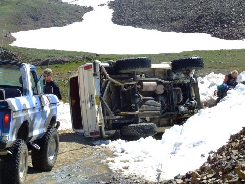 1987 Ford Bronco - A white SUV has overturned on a snowy mountain road, with a pickup truck assisting in its recovery.