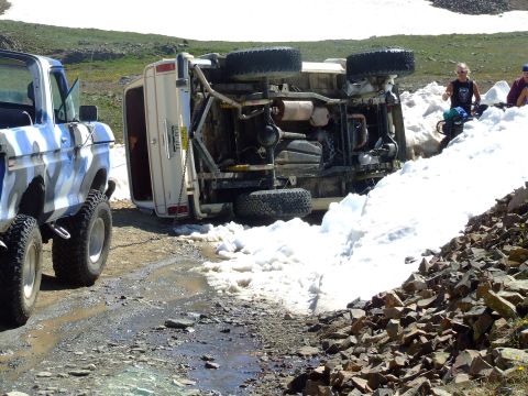 1987 Ford Bronco - A white 1987 Ford Bronco lies on its side in the snow, with a camouflage truck nearby. The scene appears to be off-road in a mountainous ar…