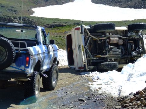1987 Ford Bronco - A camouflaged Ford Bronco stands by as another vehicle lies on its side on a snowy mountain road.