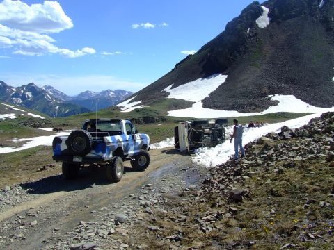 1987 Ford Bronco - An overturned vehicle lies on a snowy mountain road next to an upright, camouflaged truck.