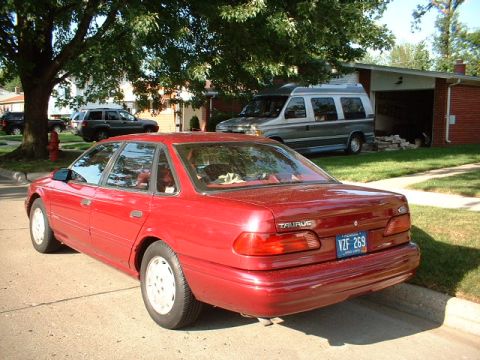 1993 Ford Taurus - Rear view of a red 1993 Ford Taurus sedan, with other vehicles visible in the driveway and street.