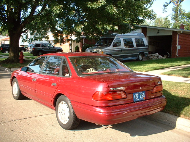 Red 1993 Ford Taurus sedan parked on a street, with a silver van and a dark SUV in the background
