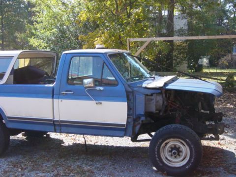 1985 Ford F-250 - This blue and white Ford pickup truck is missing its front grille and bumper, revealing exposed engine components.