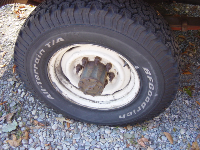 Close-up of a BFGoodrich All-Terrain T/A tire on a white rim with a rusty hub. Gravel surrounds the tire