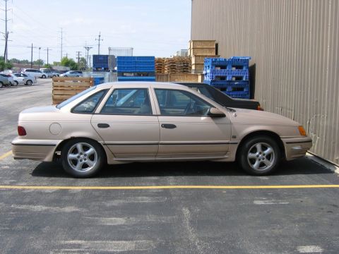 1991 Ford Taurus - Side view of a tan sedan parked in a lot with blue and brown crates stacked nearby.