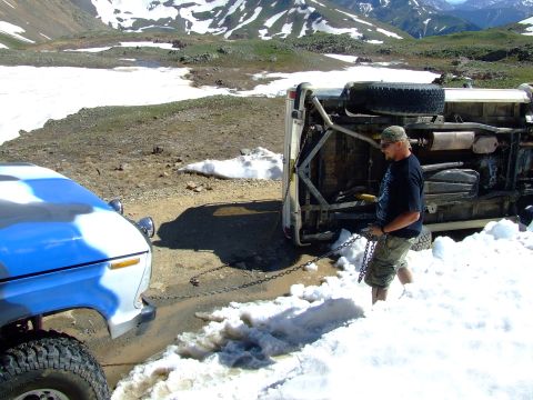 1987 Ford Bronco - A man winches a flipped SUV from a snowy mountain pass, with another vehicle assisting.