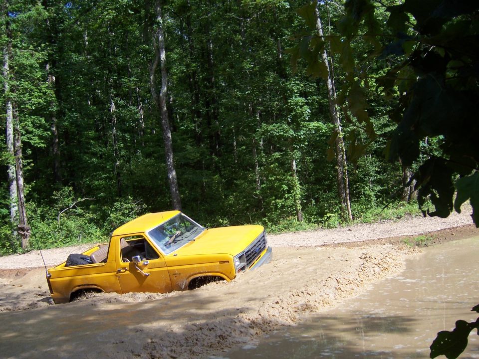 Yellow doorless early-1980s Ford Bronco driving through a deep mud pit in the woods