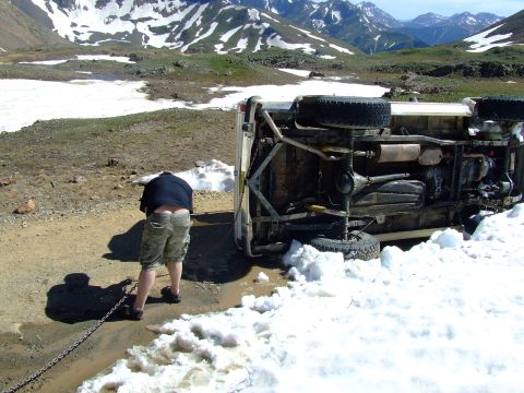 1987 Ford Bronco - A vehicle has rolled over on a dirt road with snow patches and mountains in the background.
