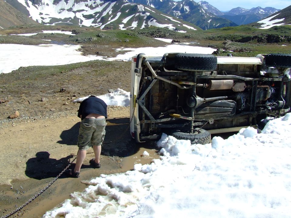 guy pulling chain attached to overturned vehicle on snowy mountain road