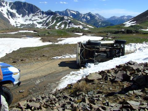1987 Ford Bronco - A white 1987 Ford Bronco lies on its side, being pulled by another vehicle on a rocky, snow-dusted mountain pass.