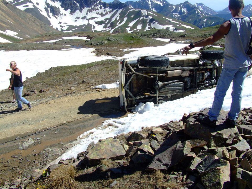 Overturned SUV on a dirt road with snow patches and mountains in background. Two people are standing nearby