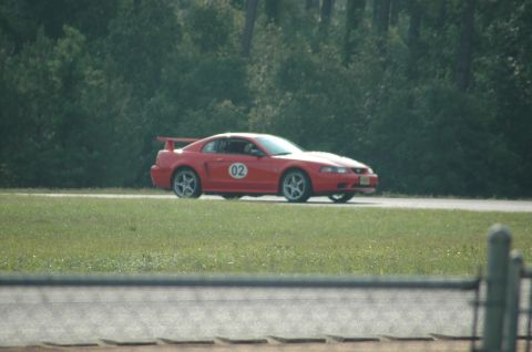 2000 Ford Mustang - A red 2000 Ford Mustang with racing decals is seen on a track, with a wooded area behind it.
