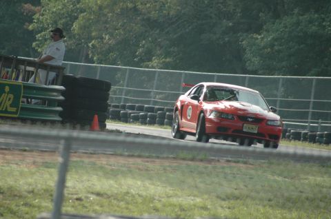 2000 Ford Mustang - A red 2000 Ford Mustang is on a race track. A person in uniform stands on an elevated platform to the left.