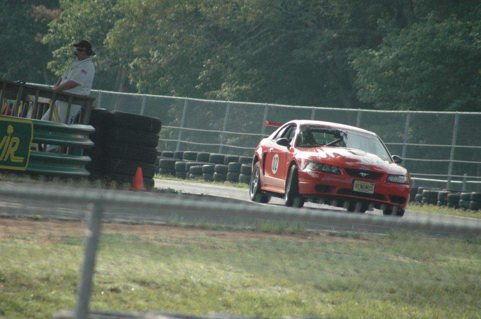 Red 2000 Ford Mustang race car on track with number 02 on door. Guy in uniform stands on elevated platform