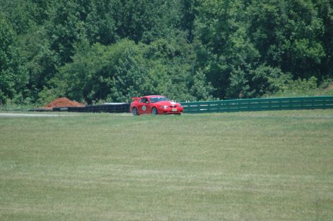 2000 Ford Mustang - A red sports car is on a race track with a green fence and trees in the background.