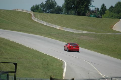 2000 Ford Mustang - A red sports car navigates a turn on a race track, with grassy hills and trees in the background.