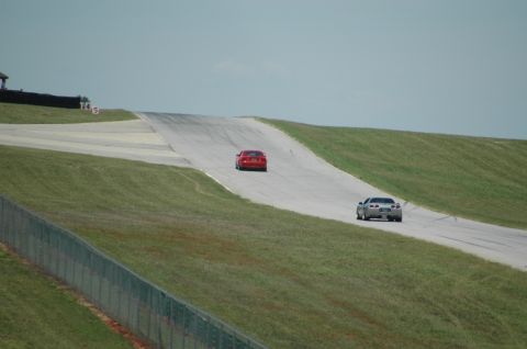 2000 Ford Mustang - Two sports cars race on a track with grassy hillsides under a clear sky.