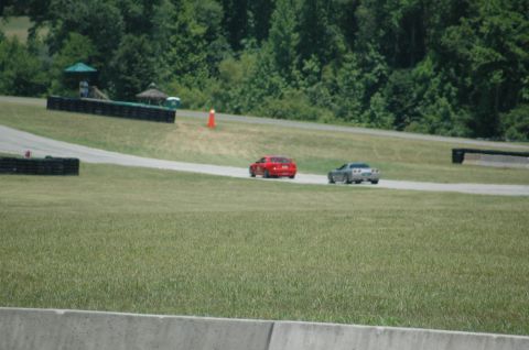 2000 Ford Mustang - Two sports cars race on a track with trees in the background.