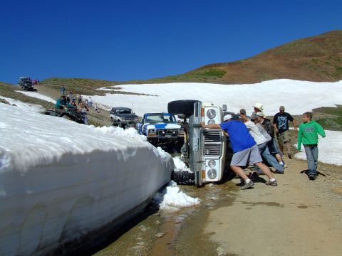 1987 Ford Bronco - A group of people attempt to right an overturned 1987 Ford Bronco on a snow-covered mountain road.