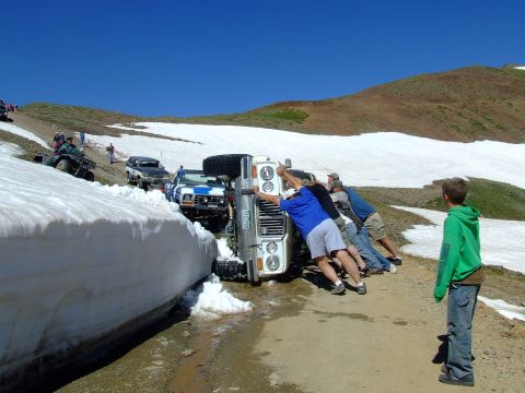 1987 Ford Bronco - People attempt to right an overturned 1987 Ford Bronco on a snowy mountain pass, with other vehicles nearby.