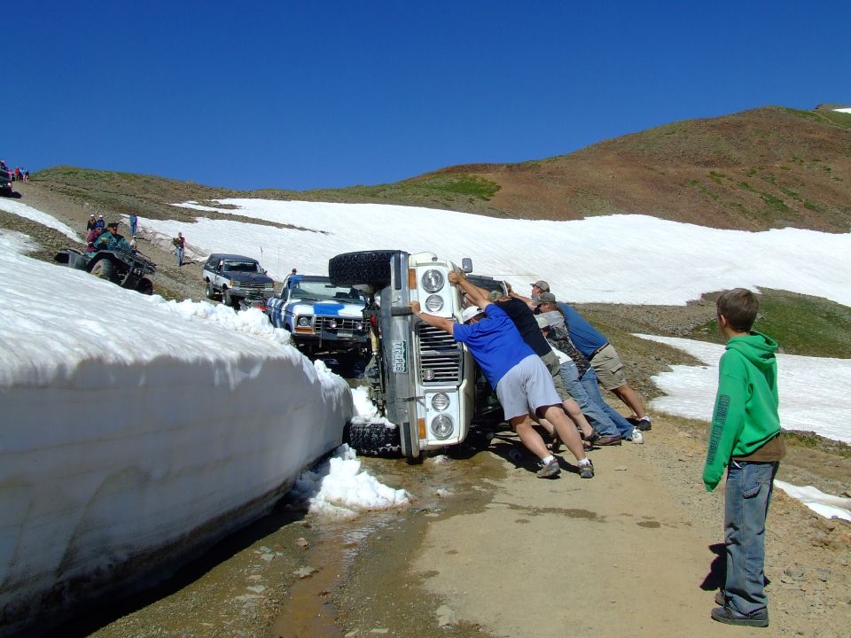 1987 Ford Bronco overturned on a mountain road, people pushing it upright. Another SUV and ATV visible