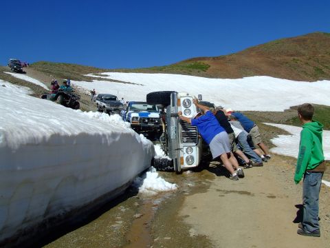 1987 Ford Bronco - A group of people attempt to right a tipped-over off-road vehicle on a snowy mountain pass.