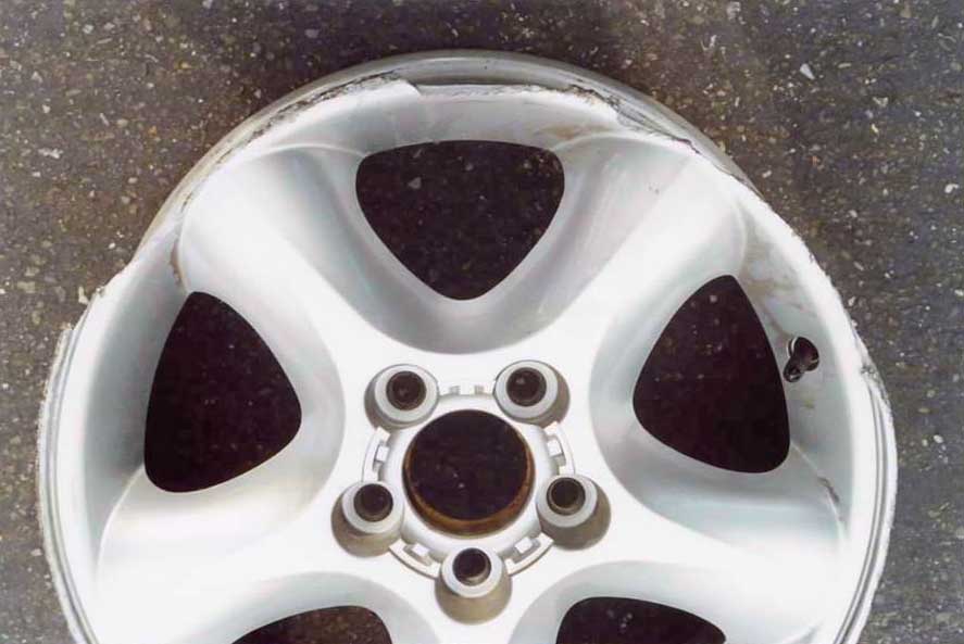 Silver 5-spoke alloy wheel without tire on asphalt, showing curb damage on the outer lip