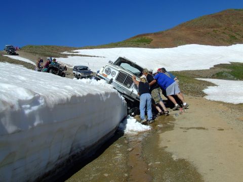 1987 Ford Bronco - People push a white Jeep Wrangler stuck in a snowbank on a mountain road.
