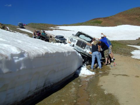 1987 Ford Bronco - Several people attempt to push a white Jeep Wrangler out of a snowbank on a mountain road.