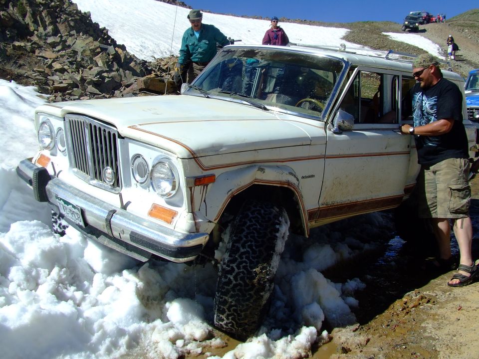 White Jeep Grand Wagoneer stuck in snow on a mountain road, people nearby