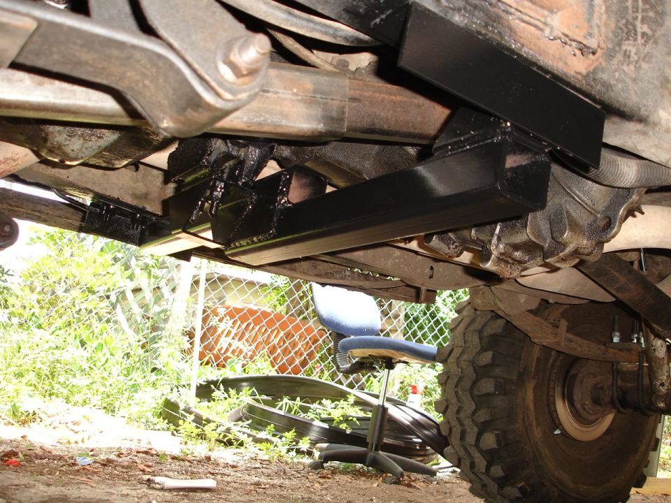Underbody of a vehicle showing a black aftermarket crossmember installed on the frame, with a large tire visible