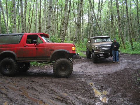 1989 Ford Bronco - Two lifted Ford Broncos are parked in a muddy forest area, one red and one tan, with a man standing by the tan vehicle.