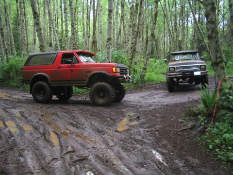 1989 Ford Bronco - Two modified SUVs navigate a challenging muddy path through a dense forest.
