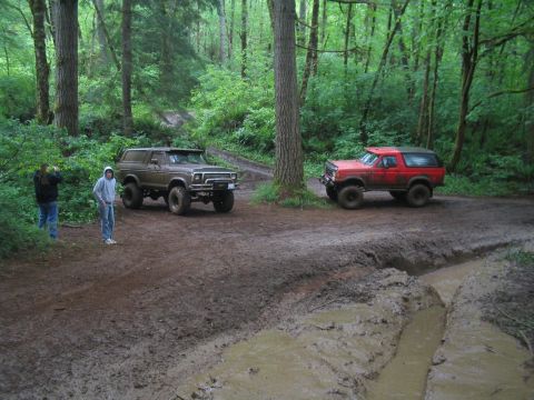 1989 Ford Bronco - Two Ford Broncos navigate a muddy off-road trail surrounded by dense forest.