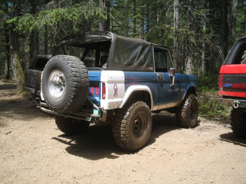 1989 Ford Bronco - A blue Ford Bronco is parked off-road in a wooded area, with another vehicle visible to its right.