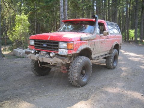 1989 Ford Bronco - A mud-covered red Ford Bronco is parked on a dirt road in a forest.