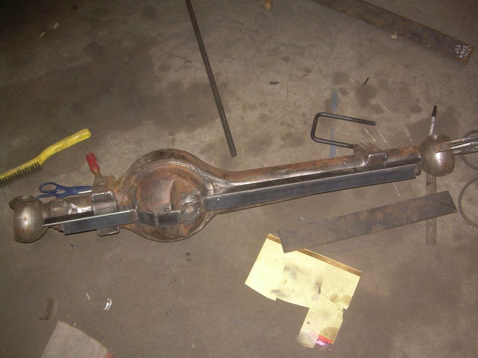 Rusty rear axle assembly with metal brackets welded on, lying on a concrete floor with tools