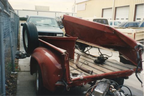 1967 Ford F-100 - A red pickup truck bed is shown with its tailgate open, revealing wooden planks and a spare tire. Another truck is visible in the backgroun…