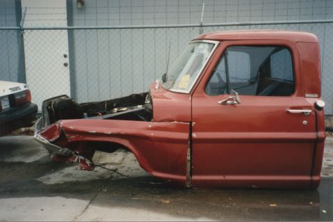 1967 Ford F-100 - The side of a red 1967 Ford F-100 pickup truck cab and front fender, showing significant damage to the front.
