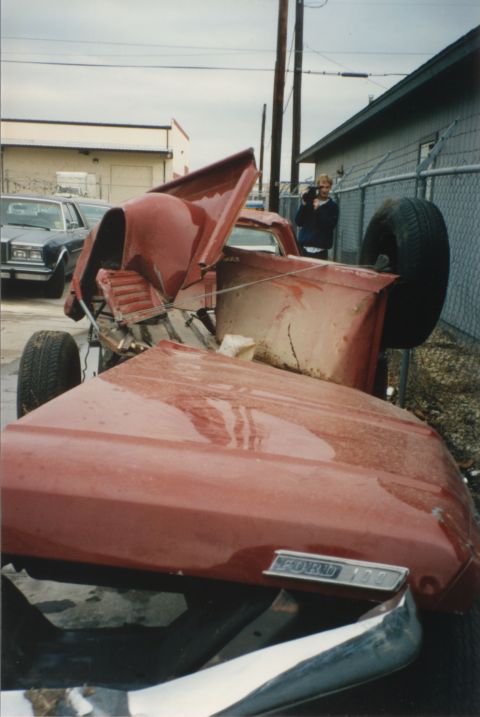 1967 Ford F-100 - A severely damaged 1967 Ford F-100 pickup truck lies on its side, its hood propped open and front end crushed.