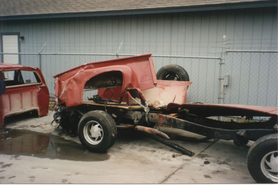 Damaged red pickup truck chassis with a separate cab in the background. The truck is missing its body panels and engine