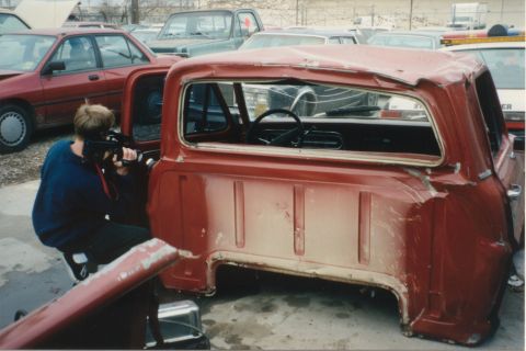 1967 Ford F-100 - A person photographs a damaged red 1967 Ford F-100 pickup truck cab in a junkyard setting.