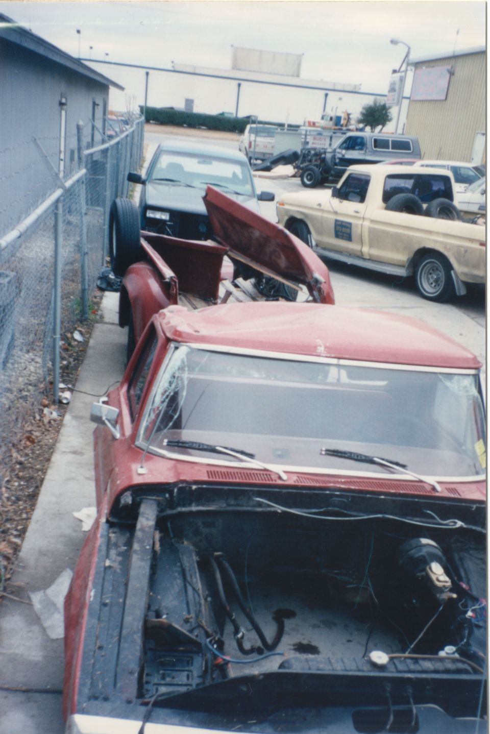 Red pickup truck with hood off, parked next to a dark pickup. Another yellow pickup is in the background