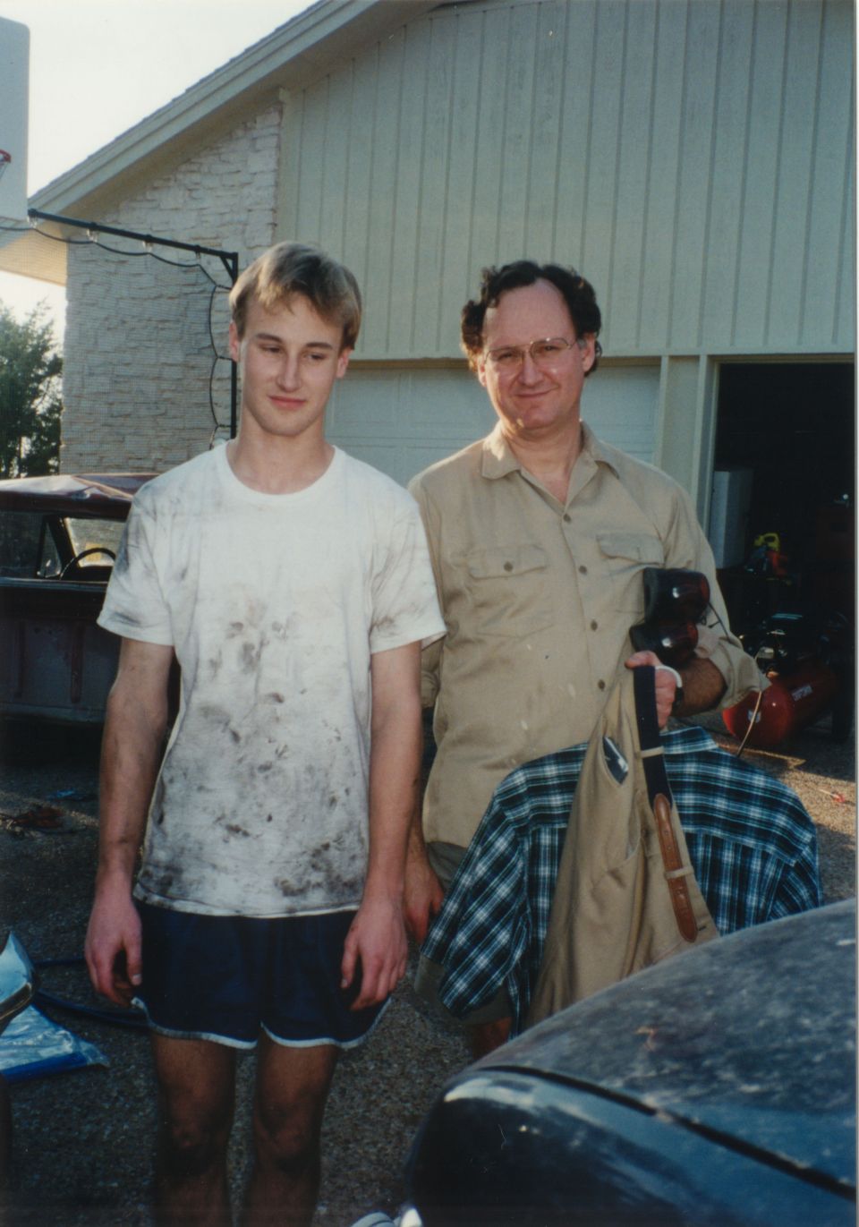 Two men, one young and one older, stand in front of a garage. The younger man wears a dirty white t-shirt and shorts. The older man holds a bag and a plaid shirt