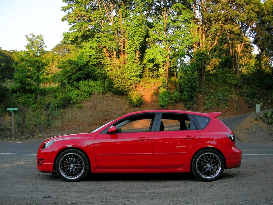 Red Mazda 3 hatchback side view on gravel roadside with gunmetal mesh wheels