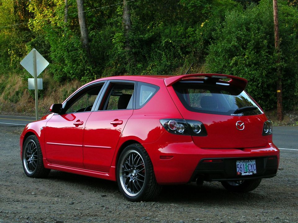 Red Mazda 3 hatchback rear three-quarter with black aftermarket wheels on gravel shoulder