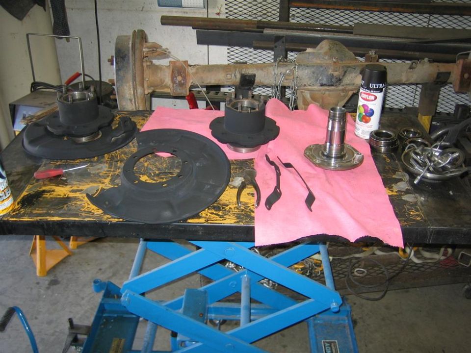 Front axle components, including hubs, brake dust shields, and bearings, laid out on a workbench with tools and a can of spray paint